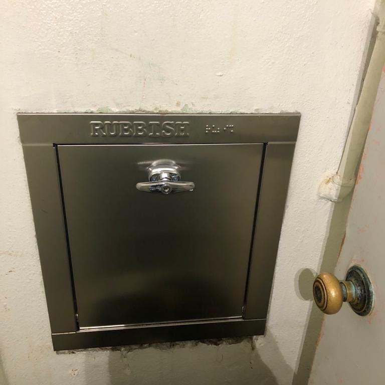 A stainless steel chute intake door marked RUBBISH, set into a hallway wall